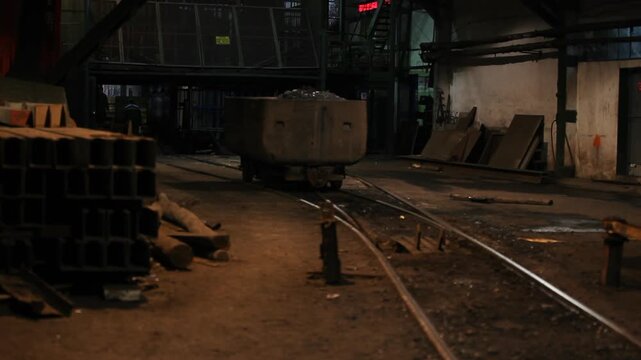 A heavy metal mine wagon filled with extracted coal or ore moves slowly along narrow railway tracks inside a dark, atmospheric underground mining facility. 