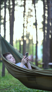 Carefree teen lounges in hammock in peaceful forest, browsing on phone. Great for summer vacation, outdoor relaxation, tech in nature and youth wellness.
