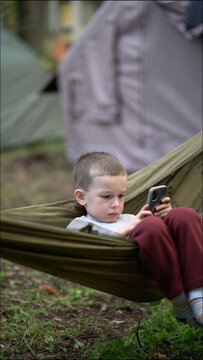 Young boy in a hammock focused on his smartphone during a family camping trip outdoors. Candid real life scene in nature with tents in the background.