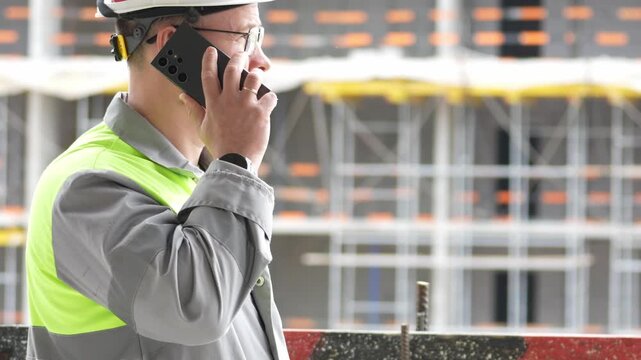 Man wears hard hat and high-visibility vest. He talks on phone at construction site. Serious expression shows focus. Scaffolding and brickwork form background