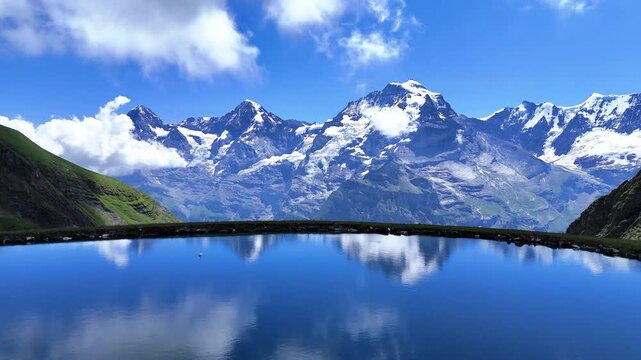 Drone view of a calm mountain lake near M&uuml;rren with snowcapped peaks and clouds mirrored on blue water in the Swiss Alps.