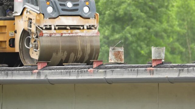 Moscow, July 04, 2026, Road roller presses asphalt on a bridge deck. Yellow machinery moves steadily. Construction worker focuses ahead. Green trees dot the background. Ideal for highway projects or