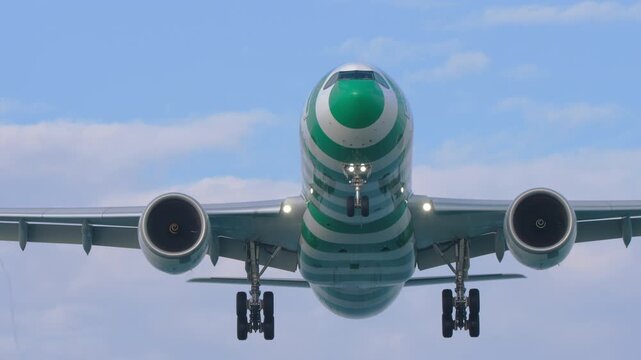 Green striped passenger plane flying low overhead for landing approach against a blue sky with clouds in daylight, low angle front view, real time