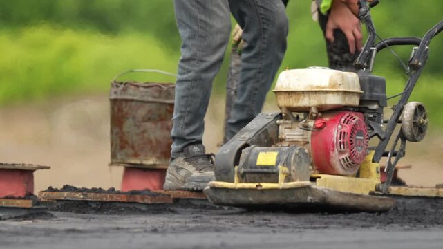 Workers operate a road roller on fresh asphalt. One wears jeans and work boots. Another wears gloves and a yellow vest. Both show concentrated effort. Rusted bucket sits nearby
