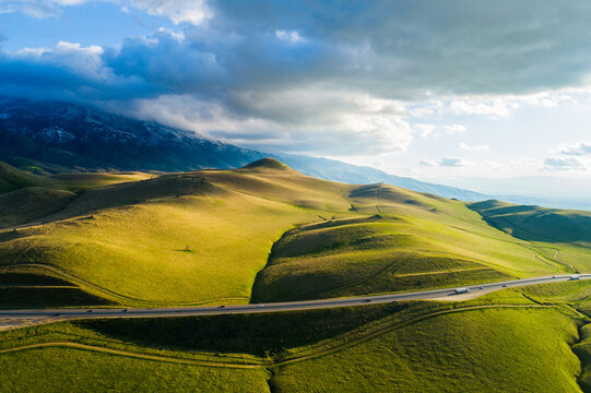 Aerial view of rolling green hills and a highway winding through the landscape with snow-capped mountains under a cloudy sky in Kern County, California, United States.