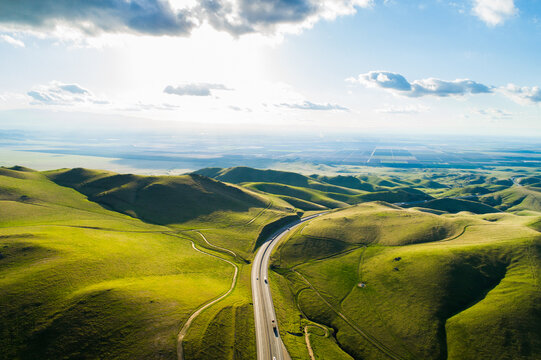 Aerial view of a highway winding through vibrant green rolling hills under a bright sunny sky in Kern County, California, United States.