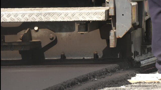 Close-up of road roller spreading asphalt. Metal tread moves steadily across the surface. Worker's legs are visible near the work area. Fresh pavement forms under the machine