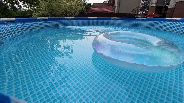 Low eye level view of round above ground pool with clear inflatable ring drifting while ripples spread across blue tiled liner in quiet backyard setting during summer daylight