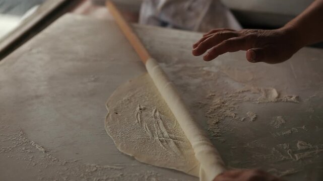 Close-up of a person's hands rolling out thin dough (yufka or phyllo) using a traditional wooden rolling pin on a floured surface. Perfect for artisan baking and authentic culinary concepts.