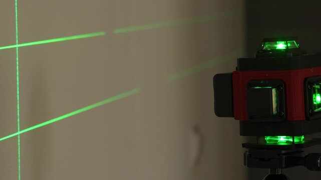 Red laser level emits bright green beams. Beams hit wall creating precise grid lines. Background shows neutral wall texture. Ideal for construction or DIY projects