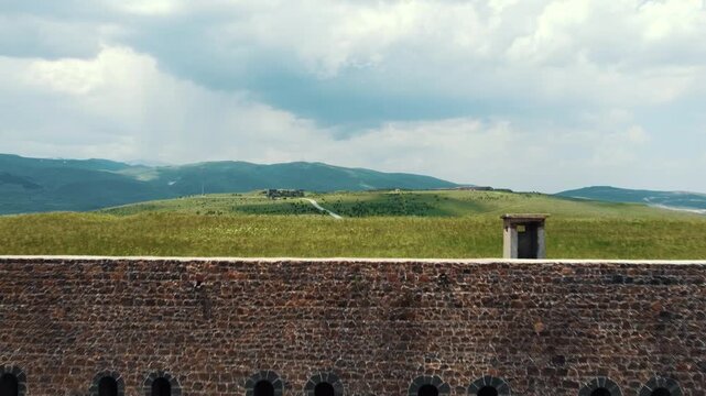 A cinematic drone shot rising over an ancient stone redoubt wall with arched windows, revealing a vast, scenic green landscape with rolling hills, a winding road, and mountains under a cloudy sky.