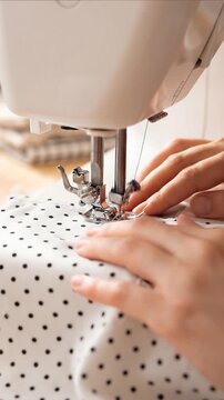 Close-up of a woman's hands sewing polka dot white fabric using a white electric sewing machine on a wooden table. Creative hobby, handmade clothes, and fashion design workspace at home.
