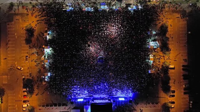 Aerial top view of crowd at night music concert. People gathered at festival outdoor stage with lights and screens, 4k