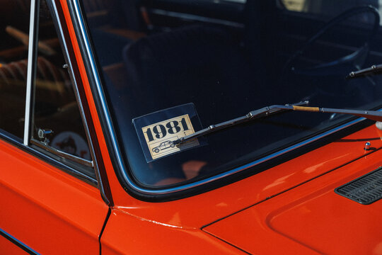 Closeup view of red classic car windshield showing aged 1981 registration sticker beneath wiper blade, chrome trim around glass, glossy orange paint, and partial interior reflection in sunlight