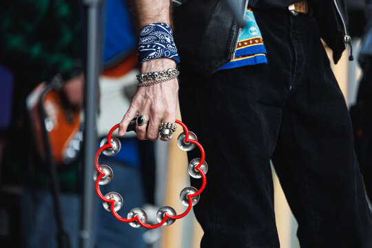Closeup view of musician left hand gripping red tambourine, with silver skull rings, chain bracelet, patterned blue bandana wrapped around wrist, leather jacket sleeve and blurred concert background