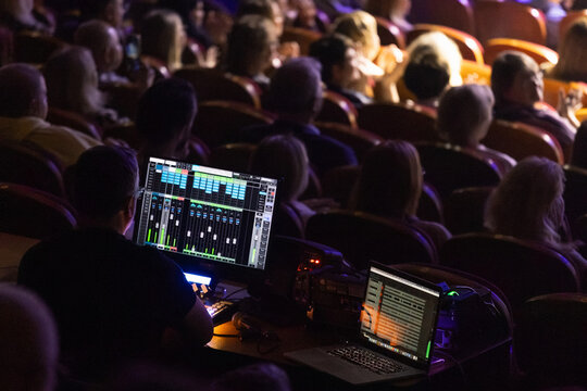 Sound engineer operates mixing console and laptop from backstage during live performance while audience sits in dimly lit auditorium; scene shows illuminated audio control screens, rows of seated