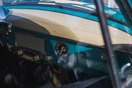 Interior view of vintage car dashboard featuring turquoise metal trim, cream padded dash top, stitched upholstery, analog clock, retro radio grille and ivory knobs in classic cabin