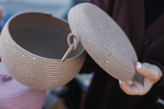 Close view of round woven storage box with lifted lid, showing coarse braided fiber texture, stitched circular pattern, decorative sequins, metal pull accent, and soft neutral styling