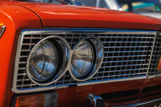 Tight front-quarter view of classic car showing chrome grille with horizontal bars, paired round headlights with reflective lenses, bright red painted hood, polished bumper details and strong contrast
