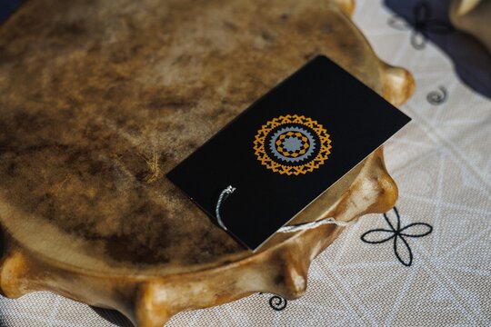 Closeup view of black rectangular gift tag featuring circular geometric gold and gray emblem resting on weathered wooden tray over textured fabric, with warm soft daylight and shallow depth of field