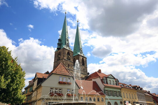 Blick &uuml;ber einen springenden Brunnen auf historische Fachwerkh&auml;user und die markanten Doppelt&uuml;rme der St. Nikolai Kirche in Quedlinburg unter einem blauen Sommerhimmel mit Wolken.