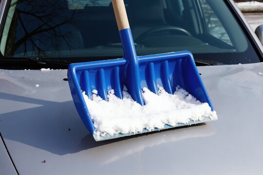 Blue shovel full of snow rests on car hood. Person clears vehicle from winter frost and ice. Cold weather preparation for safe driving on slippery roads.