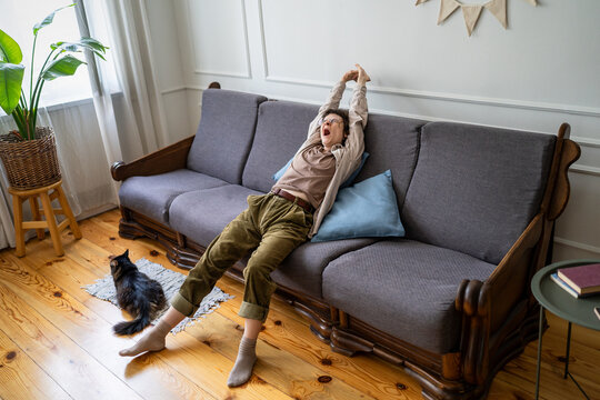 Sleepy bored woman stretching body on couch and yawning while spending lazy day at home with cat, relaxed carefree female procrastinating long hours indoors, daydreaming and resting on sofa.