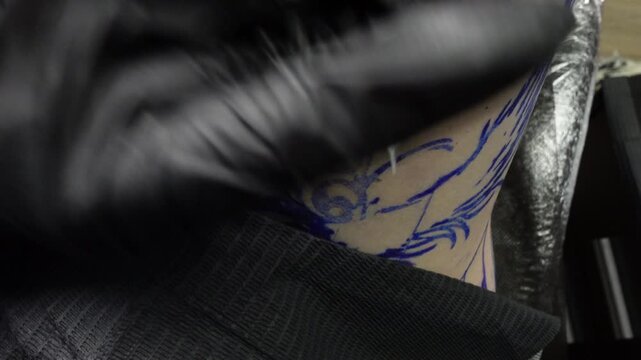 Close-up of a tattoo artist creating a black ink outline over a blue stencil during a tattoo session