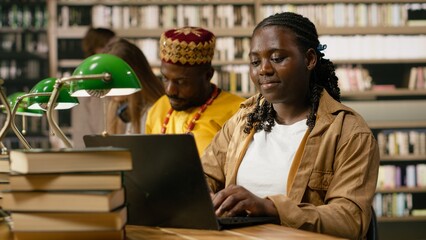Woman learner studying on laptop and typing beside stacks of books, many textbooks on shared wooden table in library. Classmates and bookshelves blurred in the background, highlighting open book. © DC Studio