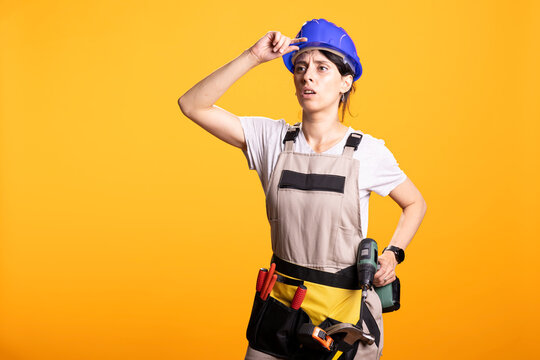 Female builder adjusts her hardhat with a puzzled expression while carrying a power drill, showing uncertainty during renovation work.
