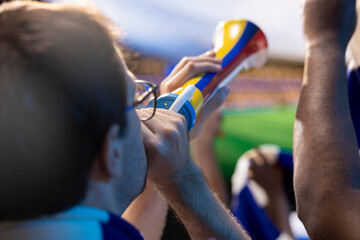 Obraz premium Soccer fans sing with vuvuzelas in stadium tribunes, blowing horns and chanting loudly to motivate their favorite football team.