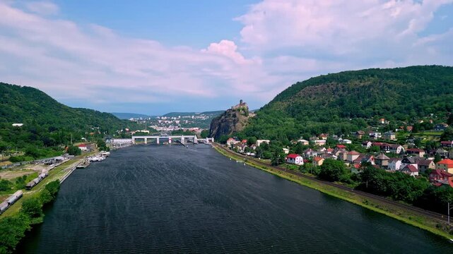 Aerial view of Strekov Castle overlooking the Elbe River, surrounded by lush green hills and residential buildings under a bright blue sky in the Czech Republic