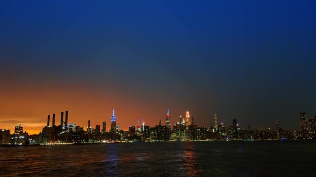Illuminated Manhattan skyline at night across the East River. City skyscrapers lit up under dark blue twilight sky with water reflection.