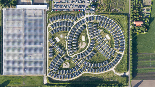 Aerial view of a modern residential neighborhood with a unique curved layout, adjacent to a large glass greenhouse complex and surrounding agricultural fields in Venlo, Limburg, Netherlands.