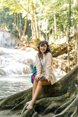Fototapeta premium Young Asian woman listening to music with headphones and mobile app by waterfall during tropical forest travel enjoy peaceful nature therapy, relaxing wellness mindfulness eco tourism active lifestyle