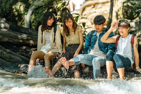Young Asian man and woman friends relaxing together by waterfall lagoon at tropical forest enjoying eco tourism adventure outdoor travel active lifestyle nature therapy on summer holiday vacation.