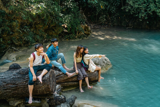 Young Asian man and woman friends relaxing together by waterfall lagoon at tropical forest enjoying eco tourism adventure outdoor travel active lifestyle nature therapy on summer holiday vacation.