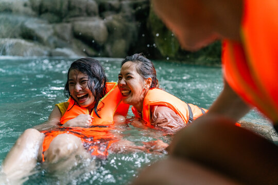 Young Asian people friends wearing life jackets and playing water together at waterfall in tropical forest, enjoying adventure outdoor active travel lifestyle nature therapy on summer holiday vacation