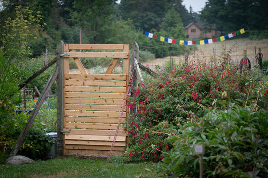 A rustic wooden garden gate leading into a cottage-style garden featuring colorful prayer flags in the background. Lovely gate helps keep deer out and adds a certain charm to the scene. 