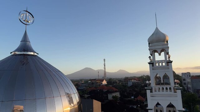 Aerial view of a beautiful white mosque minaret and reflective dome during sunrise. Cityscape with mountains in the background and clear morning sky over Indonesian town