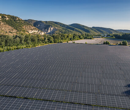 Aerial view of solar photovoltaic panels forming geometric rows at a utility-scale solar farm set against rolling hills and lush green trees in Le Pouzin, Auvergne-Rhone-Alpes, France.