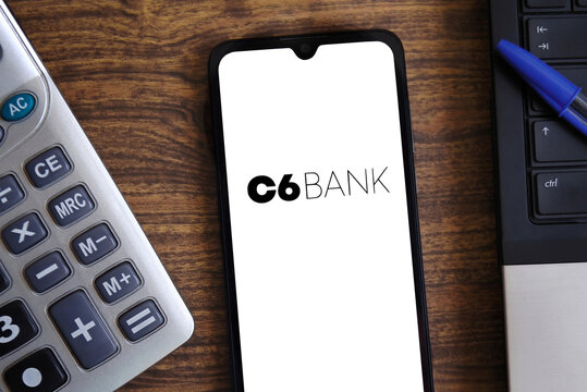 C6 Bank mobile banking app displayed on a smartphone screen next to a calculator and laptop on a wooden office desk.