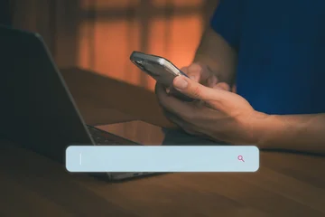 Man using smartphone and laptop with search bar for AI data browsing and SEO internet technology. Digital marketing and online information search engine concept on dark background. © ParinPIX