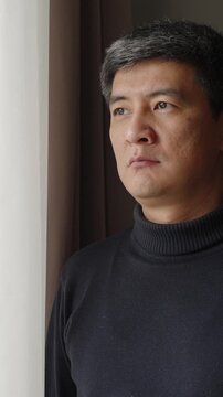 Close-up portrait of serious middle aged man looking out the window at home. Vertical shot of expressing worry, reflection, uncertainty, emotional fatigue and difficult personal decision.