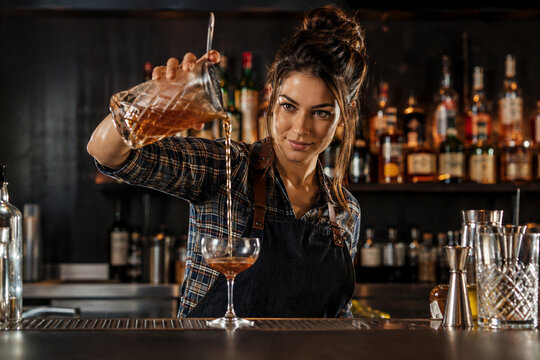 Smiling female bartender in apron pouring an artisanal cocktail into a coupe glass at a dark professional bar with copy space