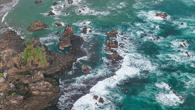 Aerial view of a rugged coastline with dark volcanic rocks, turquoise ocean waves crashing against the shore, and lush green vegetation on the cliffs in Indonesia.