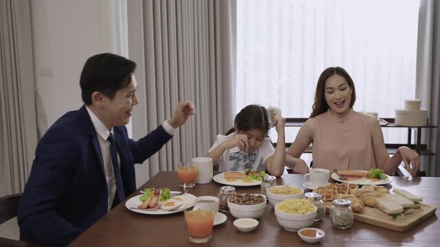 Happy Asian family father, mother, and child daughter sitting eating breakfast for health together on the dining table on cheerful morning in kitchen room at home. Dad in suit, girl in school uniform.