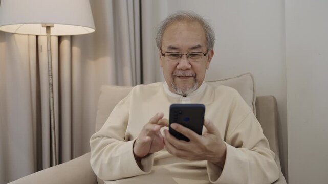 Happy Asian senior man in eyeglasses sitting smiling and using smartphone online social media, chat, shopping on sofa in living room. Old men use technology of cellphone read news and messages at home