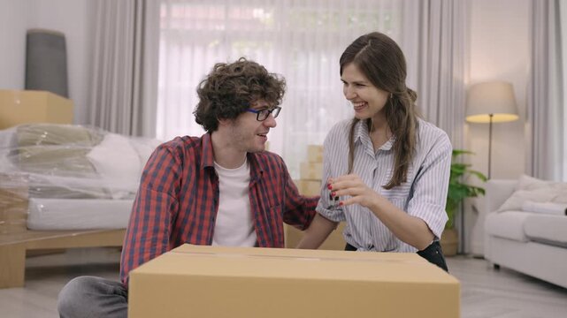 Happy young Caucasian couple relocating to new home. Handsome husband in glasses and beautiful wife sitting smiling excited and holding a house key while moving cardboard boxes and furniture indoors. 