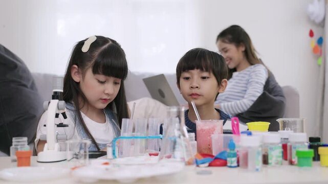 Happy adorable Asian boy and girl are sitting learning science and conducting chemistry experiments together with a young teacher sitting and taking care in the back of a nursery classroom lab. 